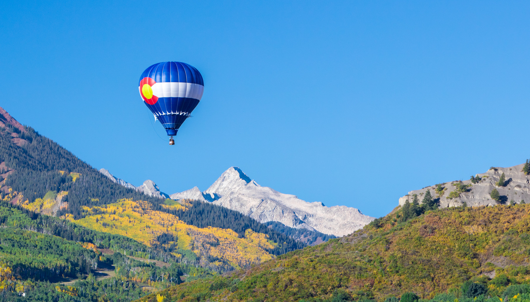 Landscape photo of hot air balloon with Colorado flag on it  above the trees during fall in Aspen, Colorado.